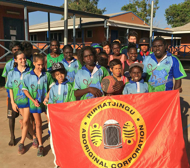 Group of young Yolngu athletes standing with a man in front of a flag of Rirratjingu Aboriginal Corporation