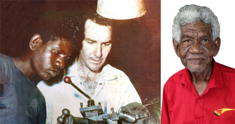 Young Aboriginal man looking at machinery with a white man, and as an older man, in a red shirt, looking at the camera