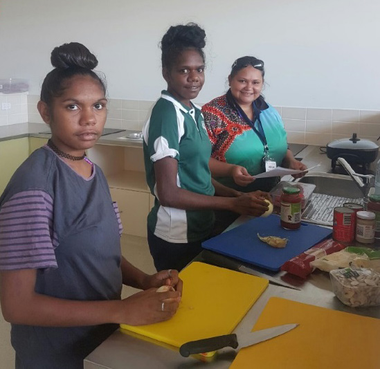Two young Aboriginal women prepare fresh food in a kitchen while a third woman looks on