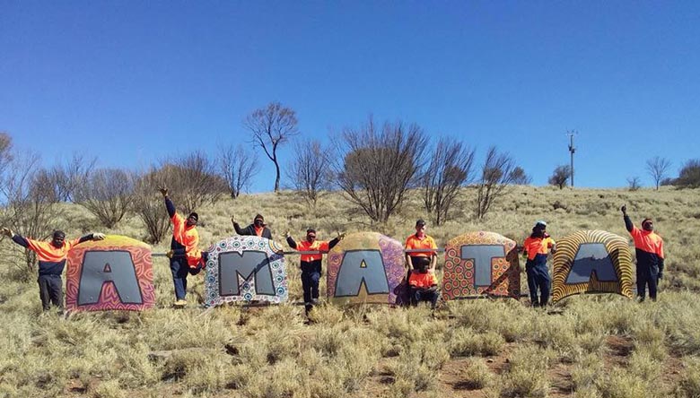 Eight Anangu people pose next to a huge sign for the community of Amata, which is made from car bonnets