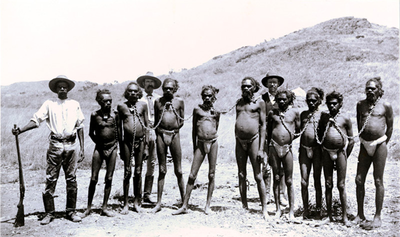 Three hatted white men, one with a shotgun, standing in front of a hill—guarding nine Aboriginal men wearing loincloths and heavy chains between their neck collars