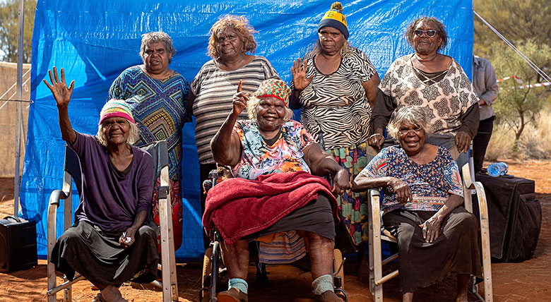 Seven Anangu women with a blue backdrop in a central desert setting