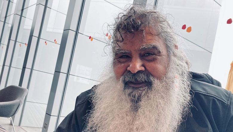 Close-up of Jeffrey Newchurch, an Aboriginal man with a long white beard, smiling in front of a glass wall