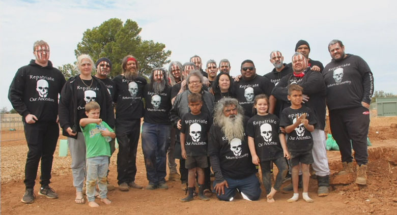 Group of 22 adults and children, many wearing a black shirt printed with a white skull and the words 'Repatriate Our Ancestors'