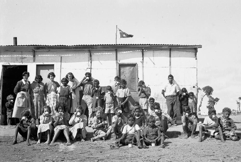 Large group of mostly Aboriginal people standing and sitting outside a metal-roofed building drinking from enamel cups. Above the building the Australian flag is flying