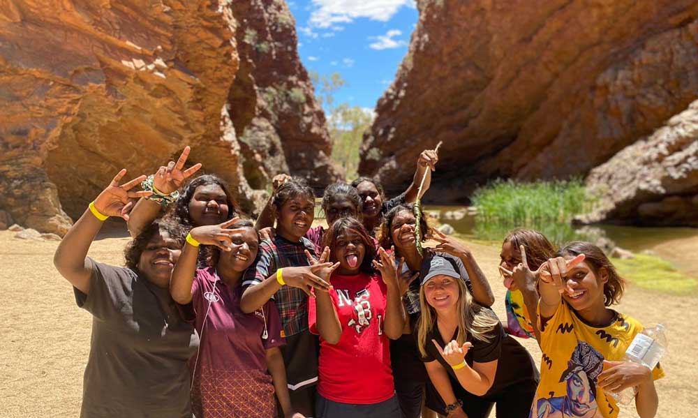 Group of 9 young women from Arlparra, 8 of them Aboriginal, standing proud and happy, close together, posing for the photo in front of a canyon