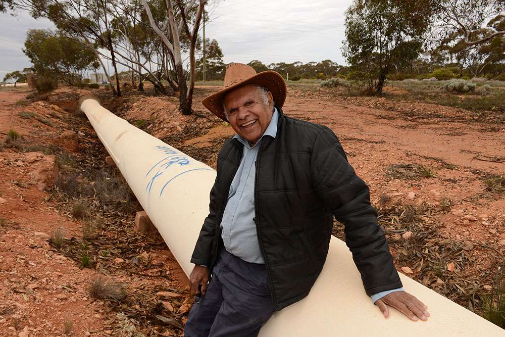 Brian Champion, Aboriginal director of Goldfields Aboriginal Languages Centre Aboriginal Corporation, leaning against a concrete pipeline on his Country, smiling at the photographer