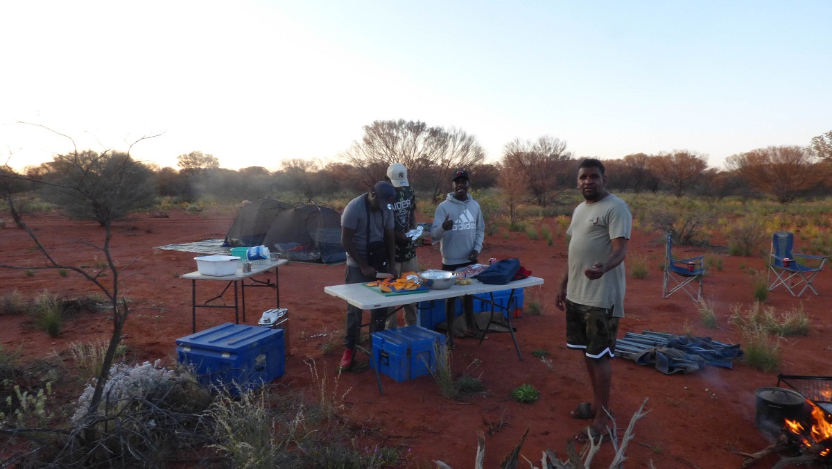 Three men stand, preparing food, at a trestle table in a bush setting with red dirt. Behind them are tents and there are camping chairs to the right of the photo. A man stands in the foreground looking at the camera