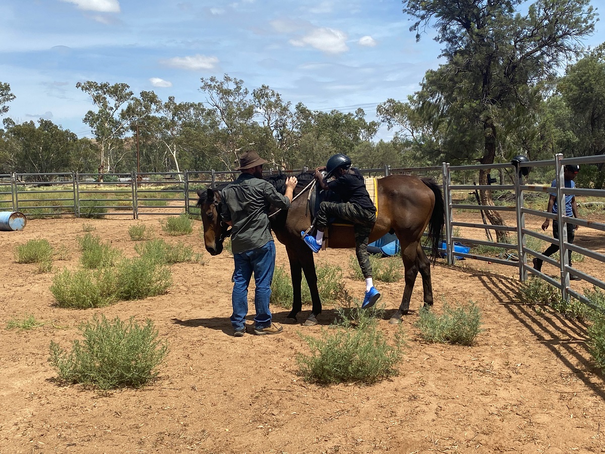 A man stands at the head of a horse holding the reins while a young person dressed in dark clothes, blue shoes and a helmet gets on. They are in a fenced yard in a bush setting