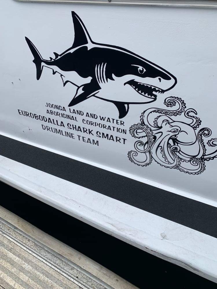 The side of a boat with an mage of a shark and an octopus and the words Joonga Land and Water Aboriginal Corporation Eurobodalla Shark SMART Drumline Team