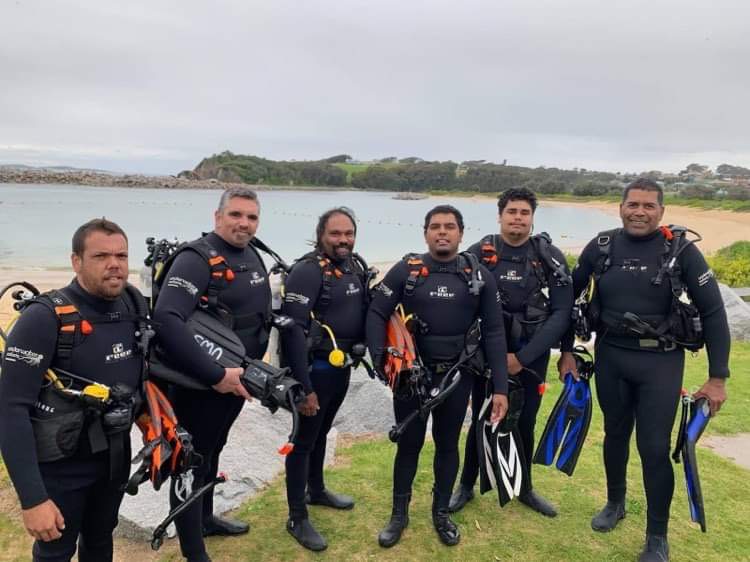 Six men wearing wetsuits and holding scuba diving gear pose with a bay and beach behind them