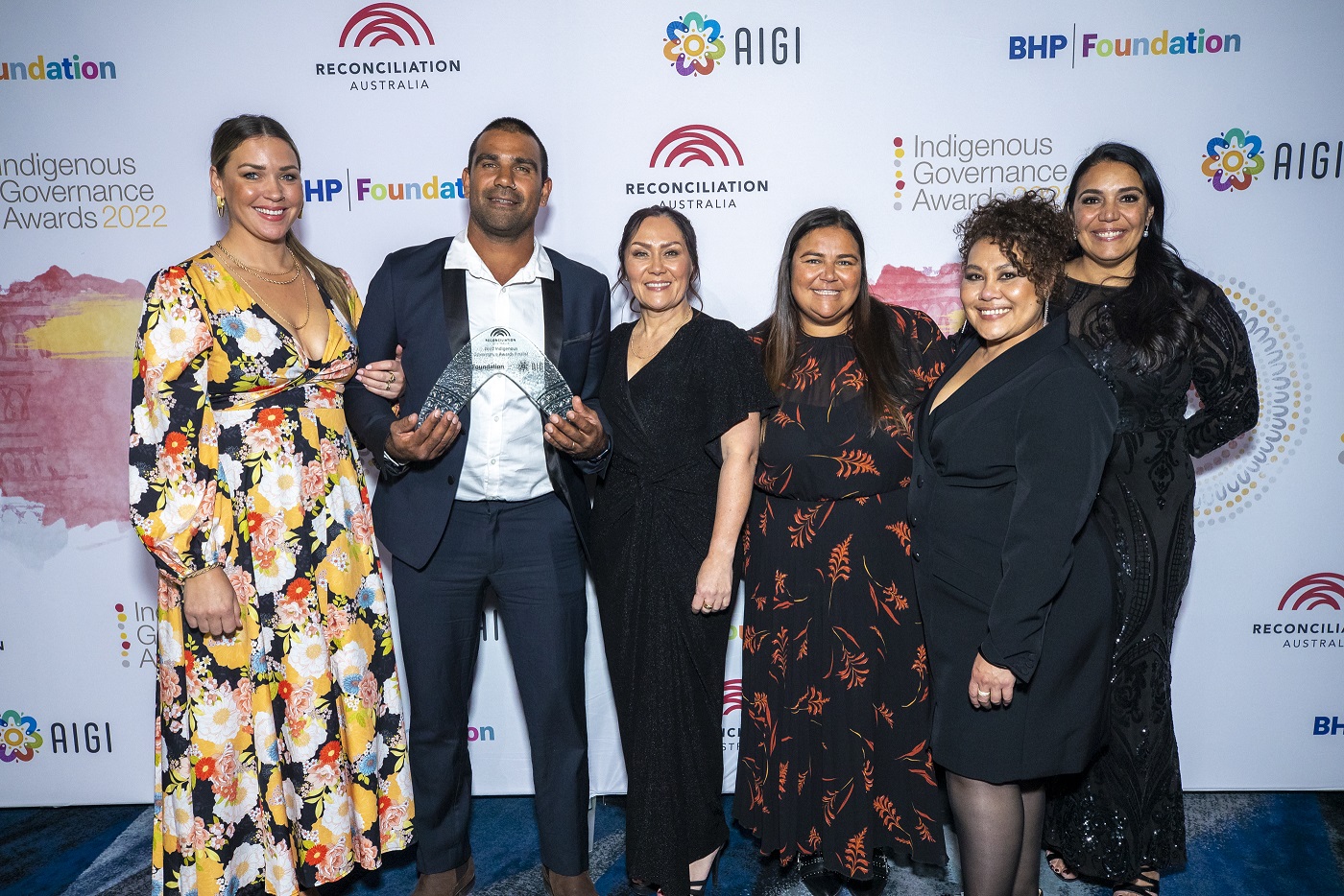 A group of people in formalwear stands in front of a backdrop with Reconciliation Australia, Indigenous Governance Awards, AIGI and BHP Foundation logos on it