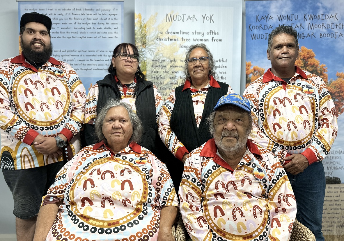 Four people stand in a row. The two middle people are women; the outer are men. In front of them sit a man and a woman. All are wearing white polo shirts that have the same Aboriginal art printed on them