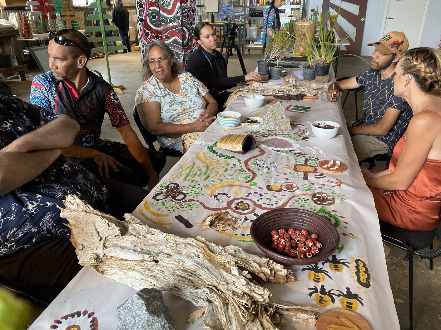 A group of Aboriginal people sit around a long table. Aboriginal artwork is painted on the paper tablecloth