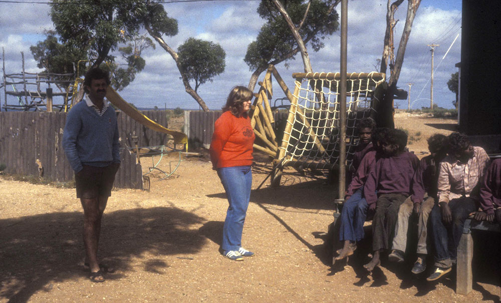 A woman stands in jeans and a red jumper in the centre of the frame. On the left is a man wearing shorts; on the right is a group of Aboriginal children seated on what could be a verandah.