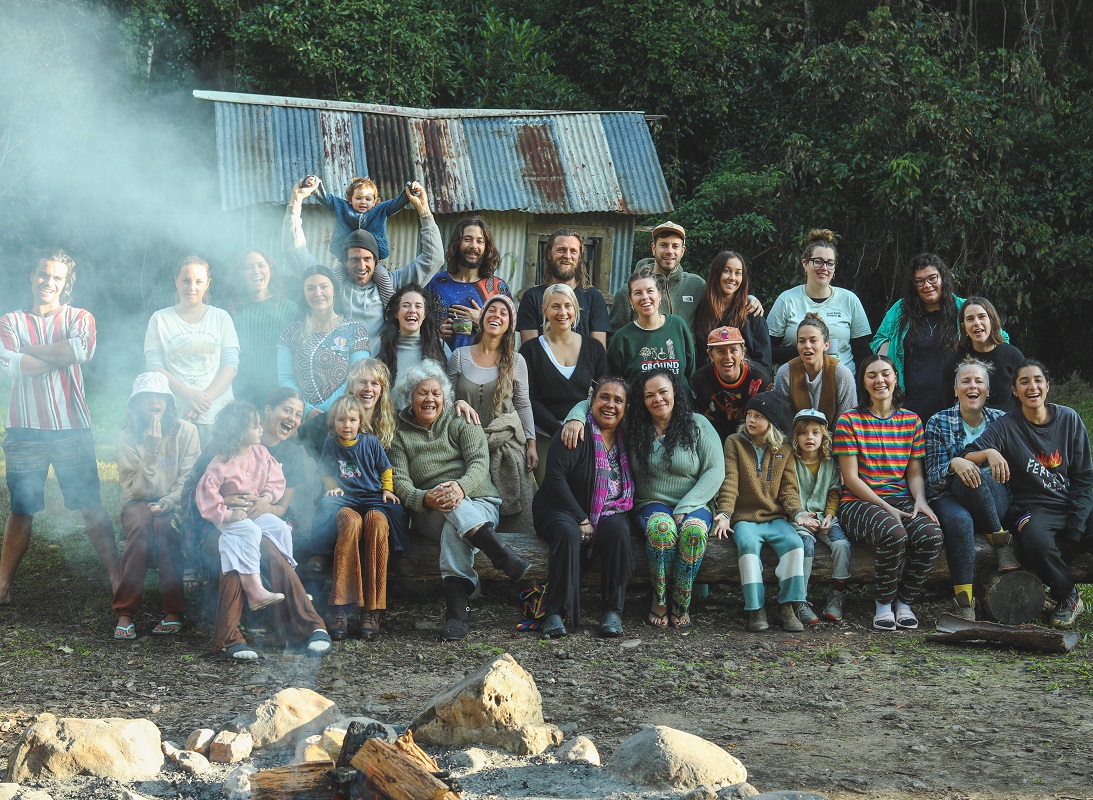 A group of people of all ages are in three rows, looking and smiling at the camera. The smoke from a campfire in the foreground obscures the faces of the people in the far left of the photo