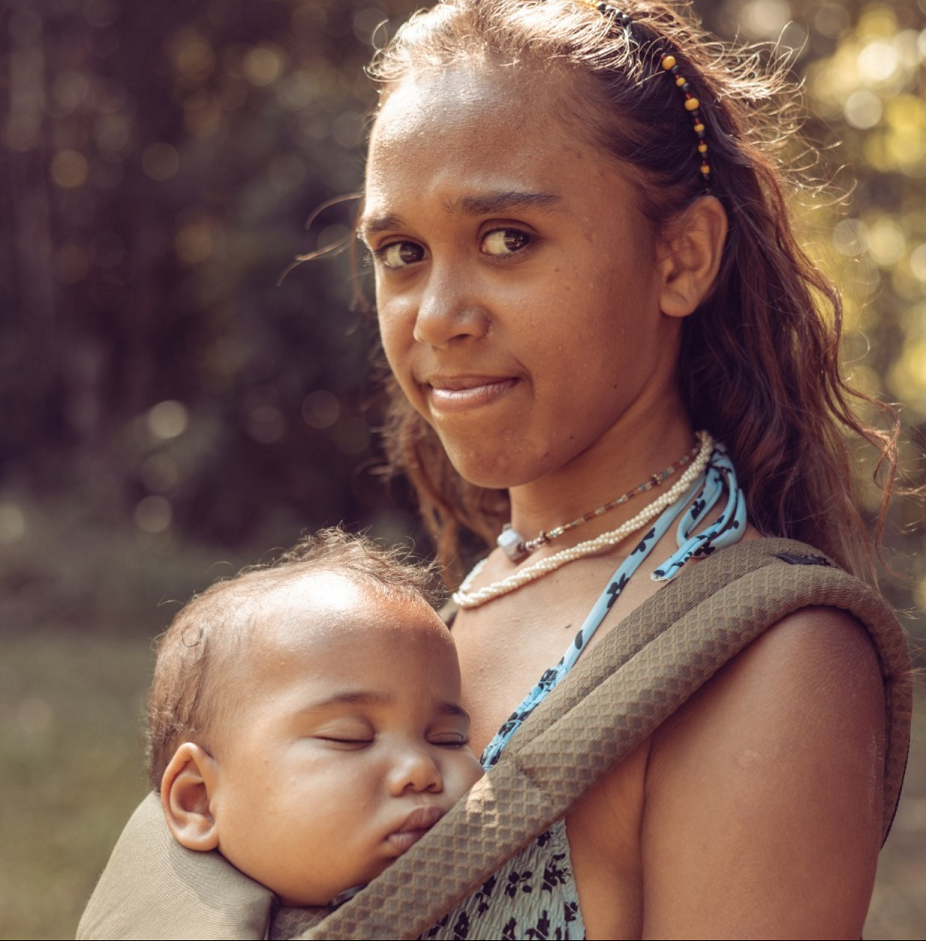 An Aboriginal woman stands side on with her head turned to look at the camera. On her front is a baby carrier with a sleeping baby inside. The baby's face is turned toward the camera