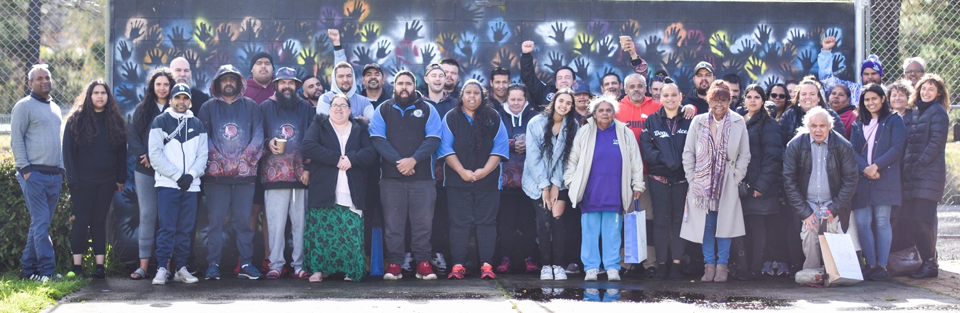 A large group of people stands in front of a dark background with handprints all over it