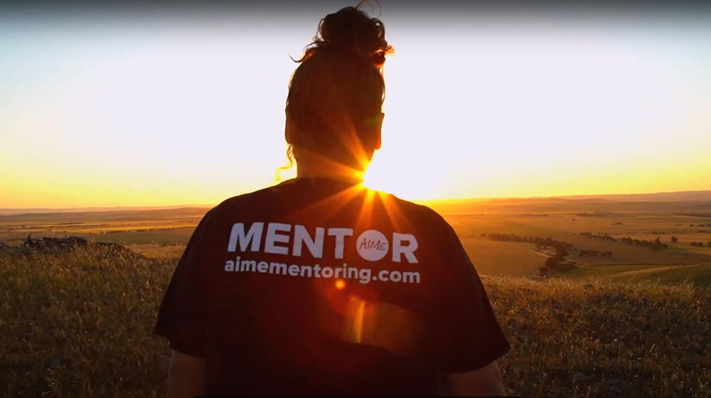 Woman standing with her back to the camera watching a sunset over open grassland. Her Tshirt is marked 'MENTOR aimementoring.com'