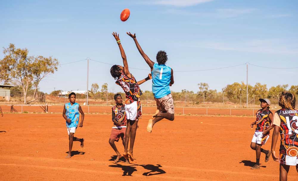 Six Aboriginal boys are visible playing football on a red dirt field. Two of them are jumping very high, reaching for the ball