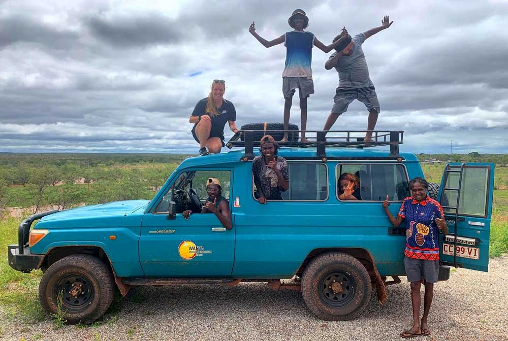Seven people in, on and in front of a land cruiser posing for the camera. Three are on the roof, 3 in the vehicle and one in front.