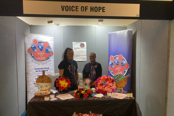 Two women stand behind a table at a stall with Voice of Hope banners on either side and above. On the table are floral wreaths and Aboriginal artwork.