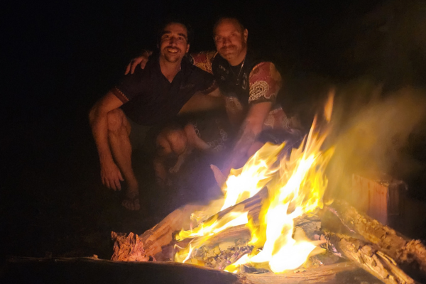 A bonfire in the foreground and 2 men sit closely together in the background