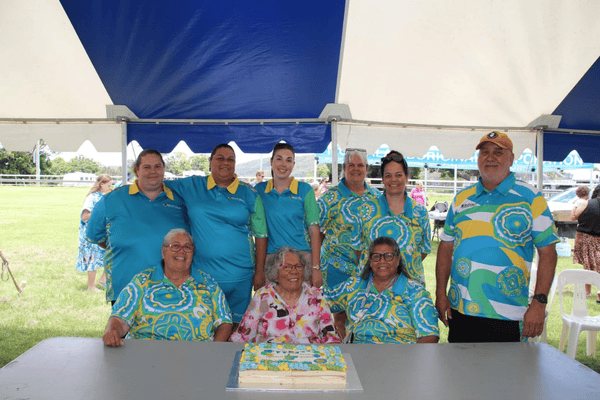 A group of people sits and stands behind a table. On the table is a decorated cake.