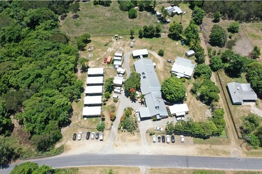 An aerial photo of buildings surrounded by trees. On the left are 5 rectangular roofs in a row, with a roadway and carpark to the right before the roof of a larger buidling and another two smaller buildings at the far right.