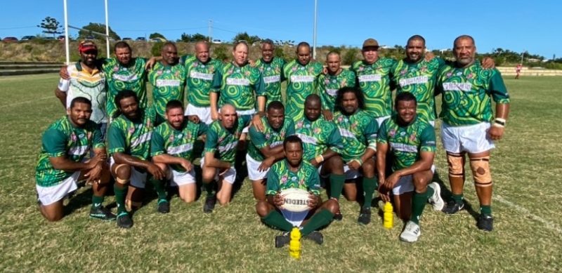 A group of men in green sporting shirts stand or kneel in two rows on a grass playing field. A man sits in front of the group holding a rugby ball