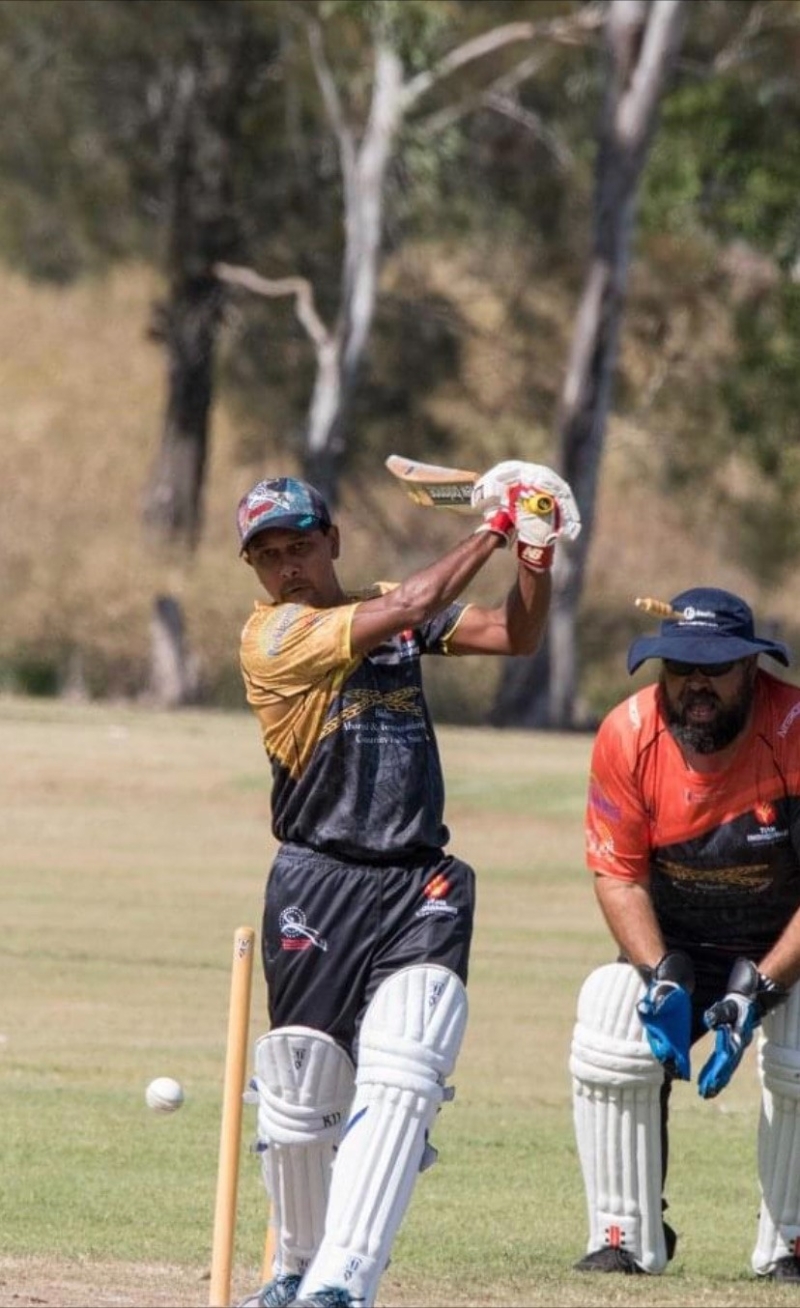 A man swings a cricket bat as a white ball goes past. To the right, a man wearing wicketkeeping gloves and pads crouches