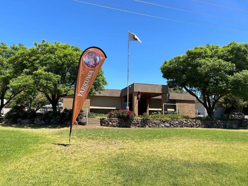 A green lawn has a a banner pole in it. The banner reads Yarkuwa. Behind the lawn is a brick building, outside of which fly the Aboriginal and Australian flags