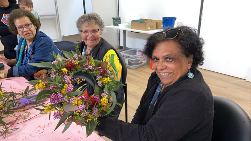 Three women sit at a table. The woman closes to the camera holds up a floral wreath.