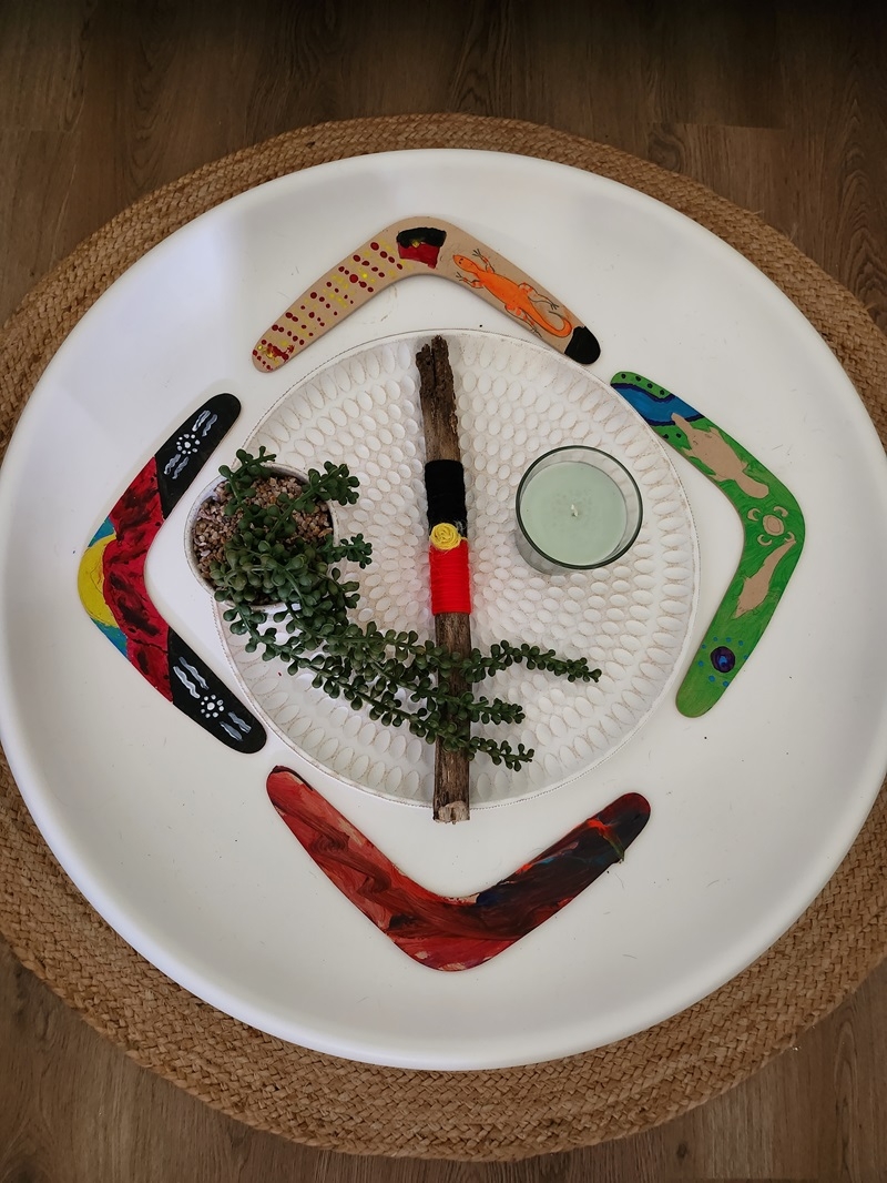 A white plate with 4 boomerangs arranged in a diamond shape. In the middle is a stick painted with the Aboriginal flag between a small plant and a candle