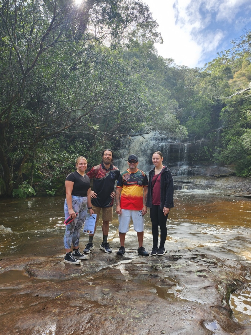 Four people stand on rocks in front of a pool of water with a waterfall in the background