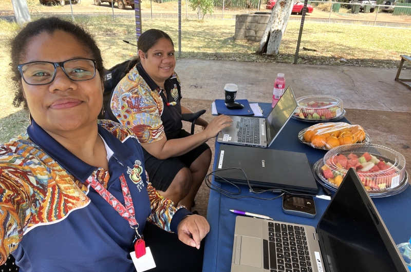 A woman holds up the camera to take a photo of herself and another woman sitting at an outdoor table with laptop computers on it