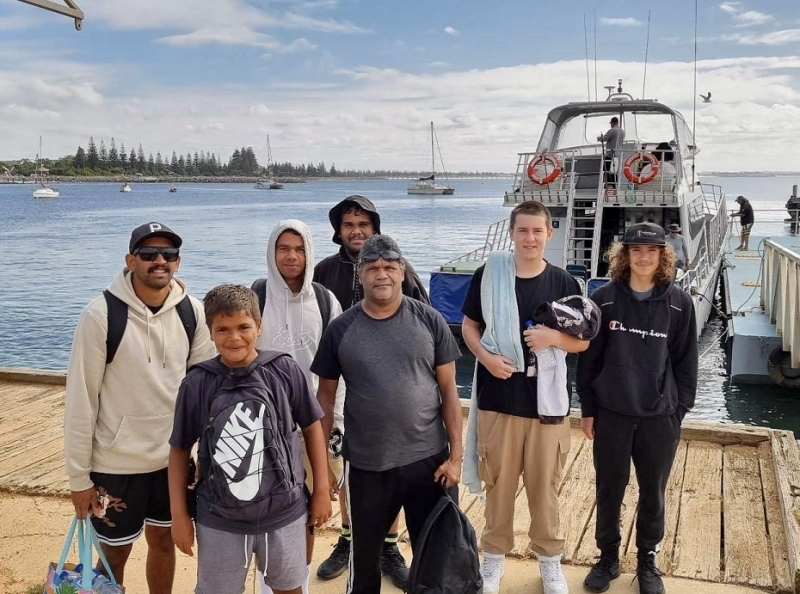 A group of people stand on a jetty with a boat behind them