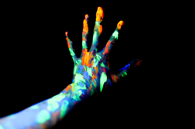 A hand covered in colourful paint glows against a black background