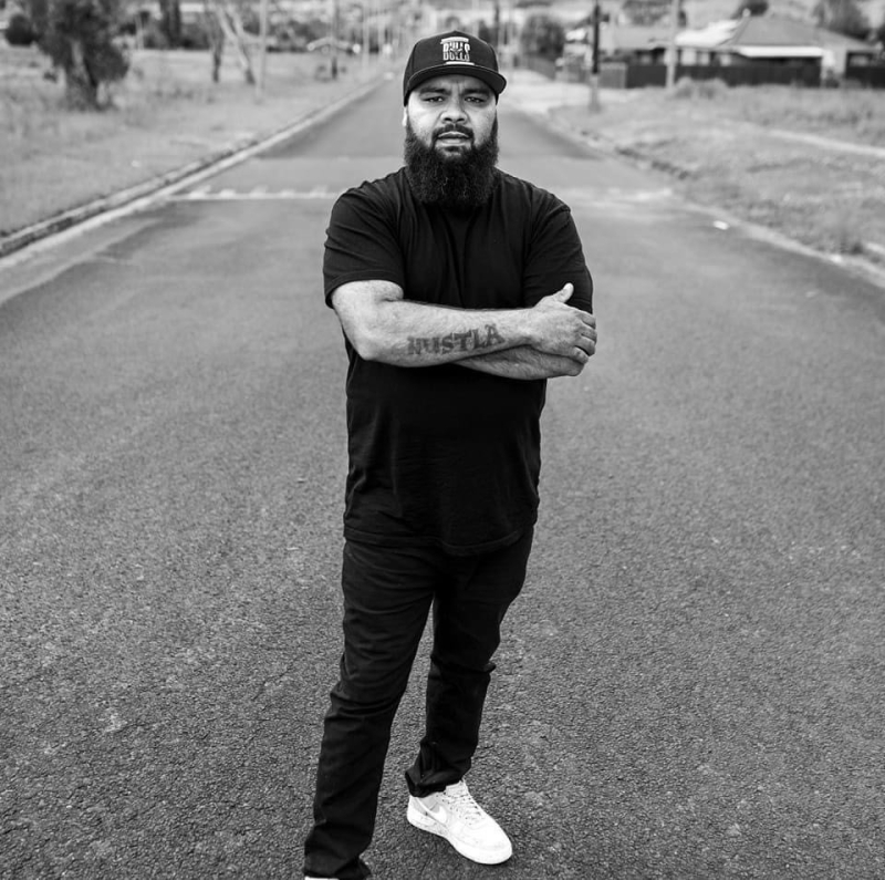 An Aboriginal man dressed in black poses standing on a road facing the camera