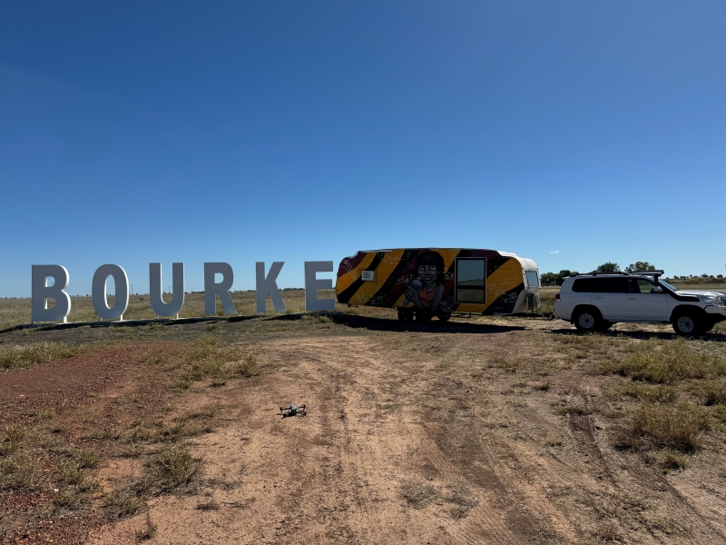 A set of large freestanding letters spells out BOURKE. Next to the letters are parked a four-wheel-drive and colourfully painted caravan