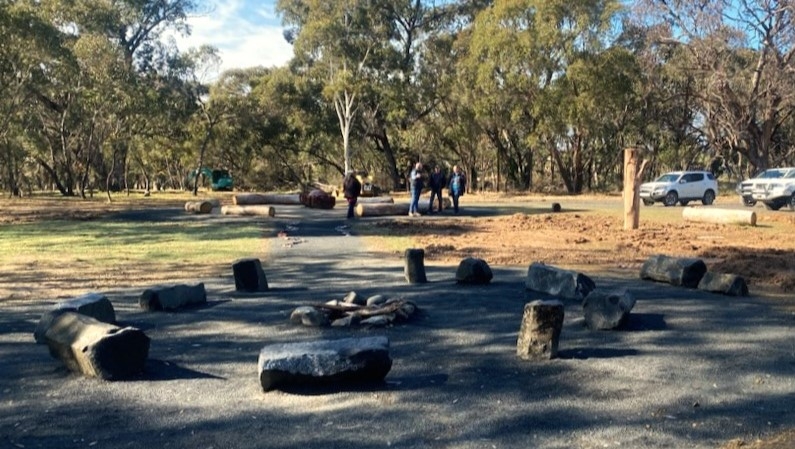 Large rocks are arranged in a circle around a stone fire pit. In the background are a group of people, a car park and large trees