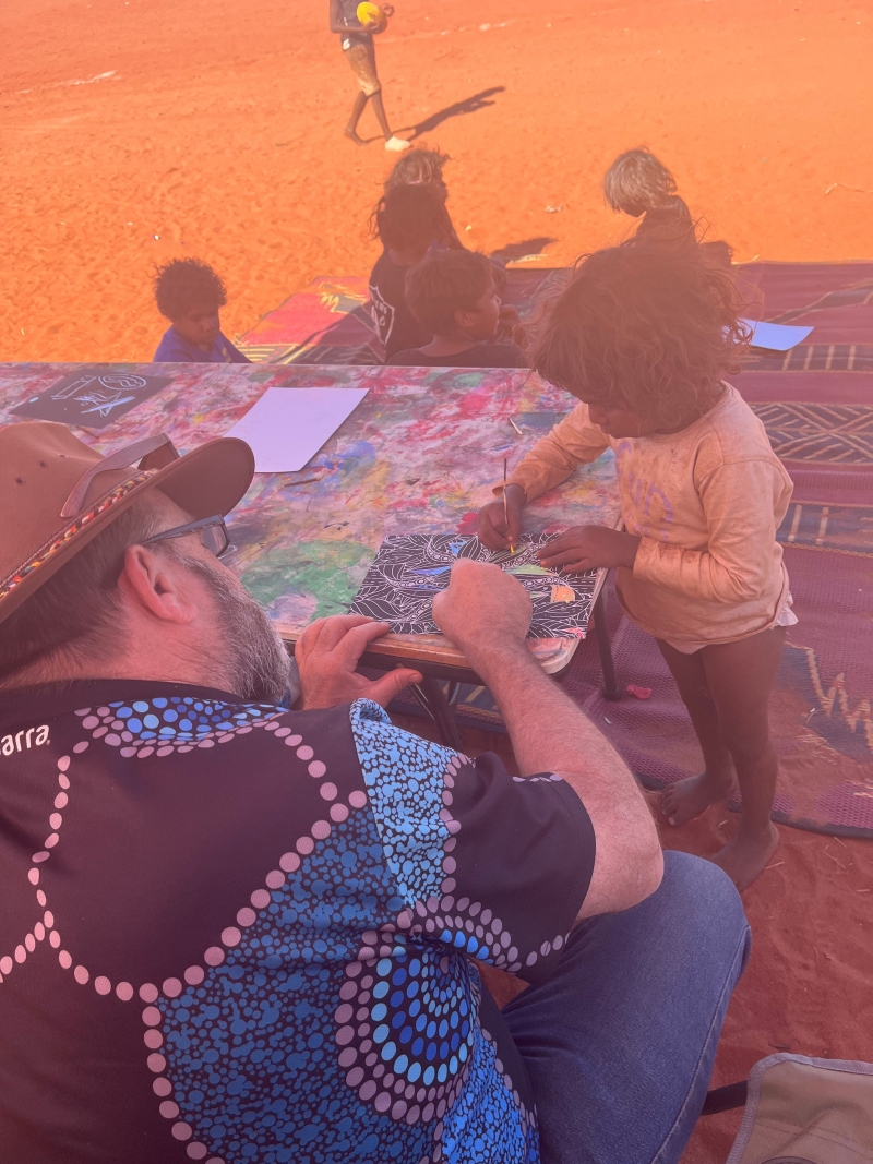 A man and a small child draw on paper at a low table. Other children and red dirt are in the background