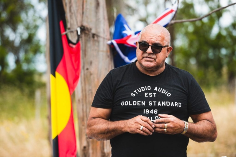 A man stands outdoors. Behind him is an Aboriginal flag