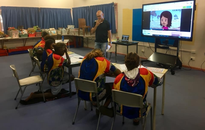 Children in school uniforms sit at desks facing a screen with an animated image on it. A teacher stands at the far side