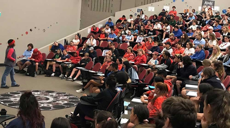 A person stands on the floor of an lecture theatre addressing people sitting