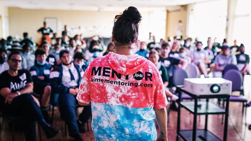 A person stands in front of a classroom of adults, her back to the camera. The back of her hoodie reads MENTOR 