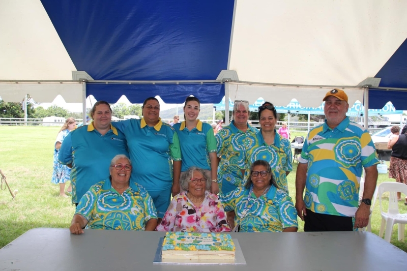 A group of 9 people stands and sits behind a table. On the table is a decorated cake