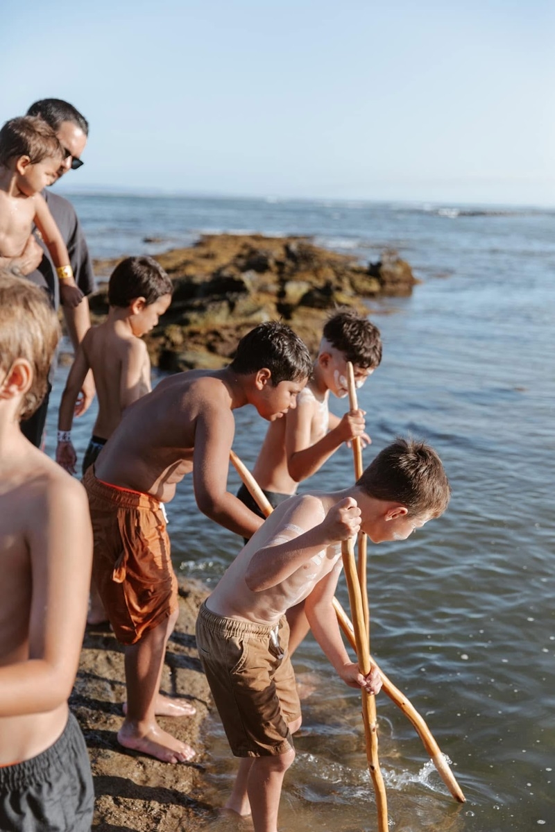 A small group of children holding spears are peering into the sea