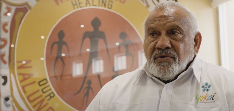 An older man with a logo on the wall behind him