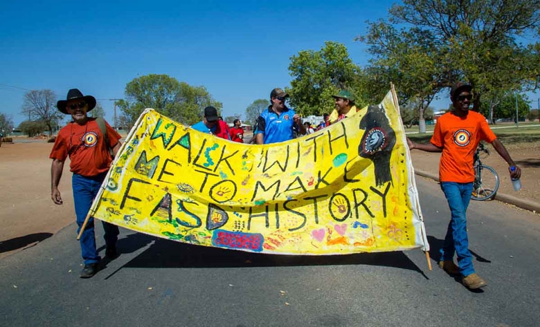 A group of people walk along a road holding in front of them a banner reading 'Walk with me to make FASD history'